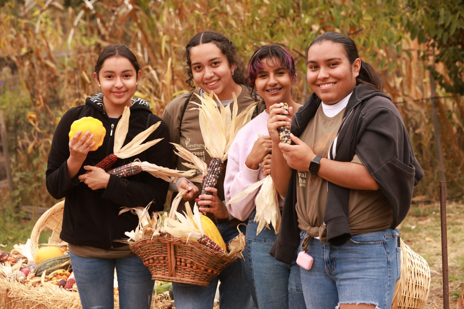 Four young people stand in front of golden corn stalks. They are smiling and hold freshly harvested squash and other vegetables from Bayer Farm. 